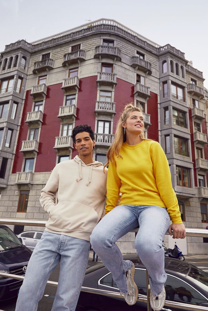 Mand, stående op ad gelænder iført en sandfarvet hoodie, kvinde i gul sweatshirt siddende på gelænder.